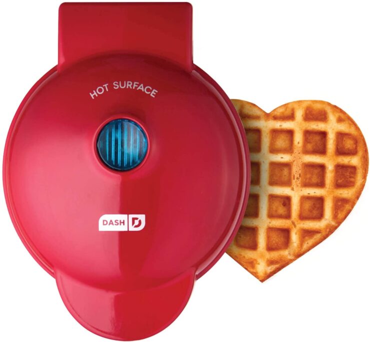 A red-colored waffle maker with a cooked heart-shaped waffle on the side. 