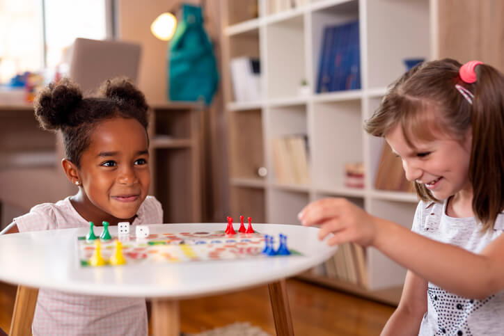 Two little girls sitting in a playroom, playing a ludo board game; one of them just won the game. Focus on the girl on the left