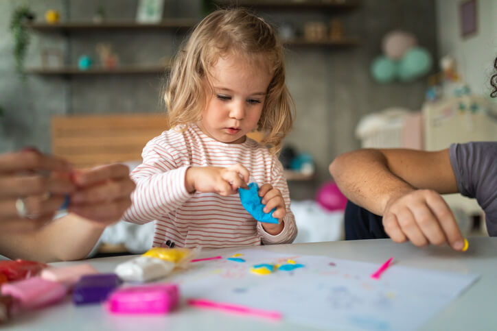 Cute toddler girl making shapes of plasticine modeling clay, unrecognizable people next to her