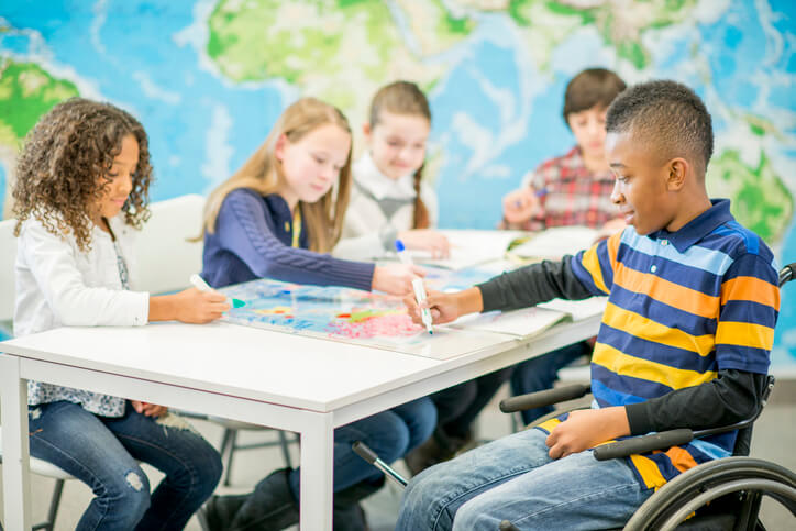 A multi-ethnic group of elementary age children are sitting at a desk and are drawing on a world map for geography class. One handicap child is sitting in a wheel chair.