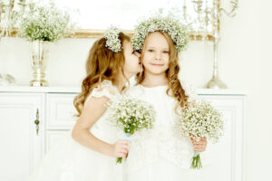 two little flower girls in wedding dresses standing at the dresser with a big mirror and held bunches of flowers